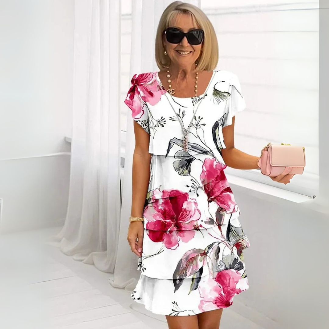 Ältere Frau in weißem Kleid mit rosa Blumenmuster, Sonnenbrille und rosa Handtasche, steht lächelnd in modernem, hellem Raum.