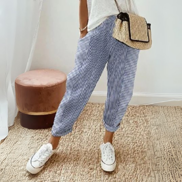 Dame in gestreifter Leinenhose und weißen Sneakers, trägt eine Strohtasche. Sommermode, lässiger Stil, bequeme Kleidung, modische Accessoires.