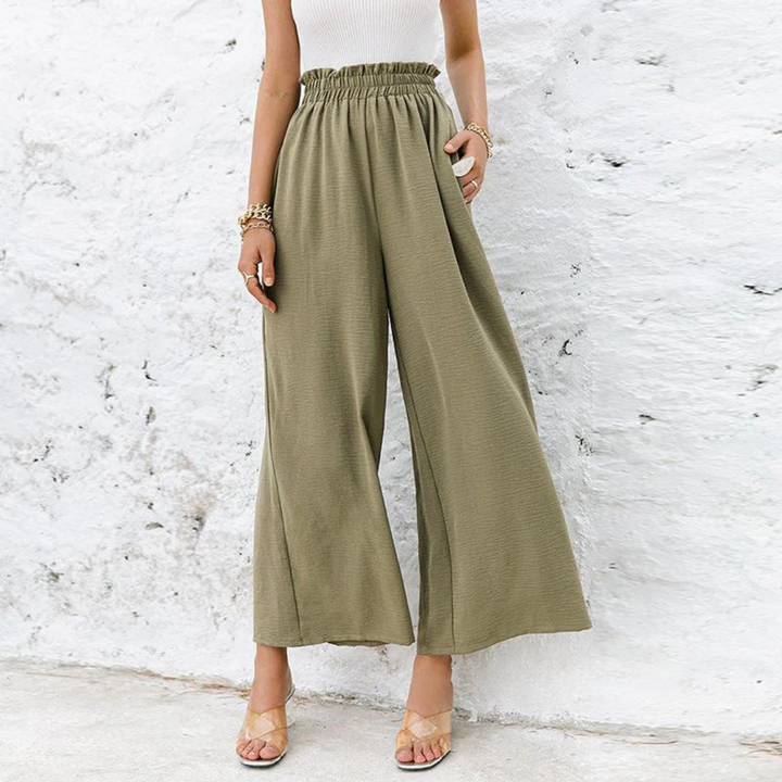 Frau in olivgrüner, weiter Hose und weißem Top vor weißer Wand. Modetrend, Sommeroutfit, Damenbekleidung, lässiger Stil.