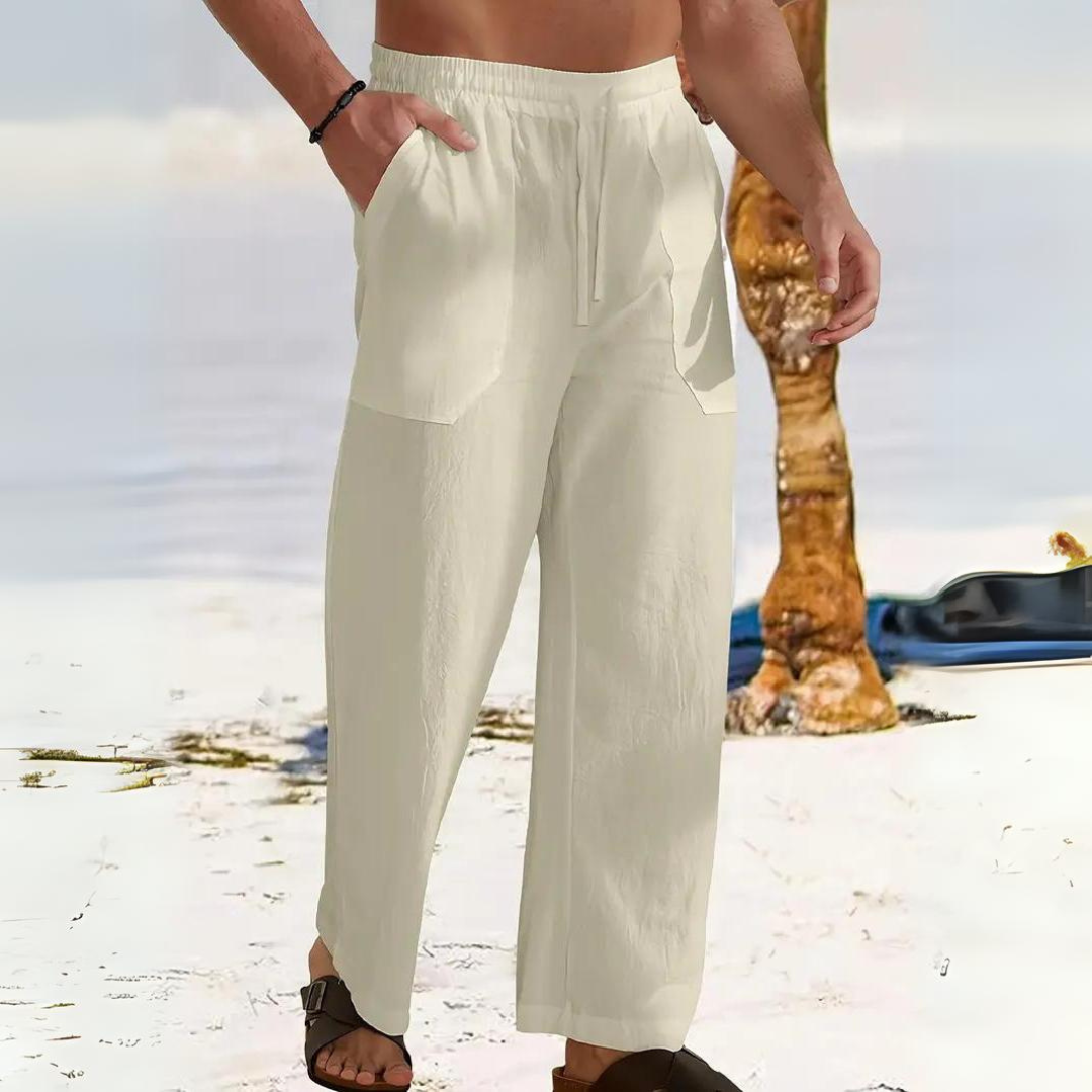 Mann in weißen Leinenhosen am Strand, Hände in den Taschen, barfuß im Sand. Sommermode, lässiger Strandlook, Herrenbekleidung.