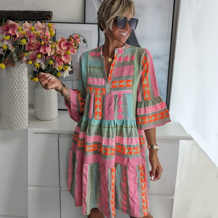 Frau in buntem, geometrisch gemustertem Kleid, steht lächelnd vor weißem Hintergrund mit Blumenstrauß. Sommermode, Damenkleid, trendiger Look.