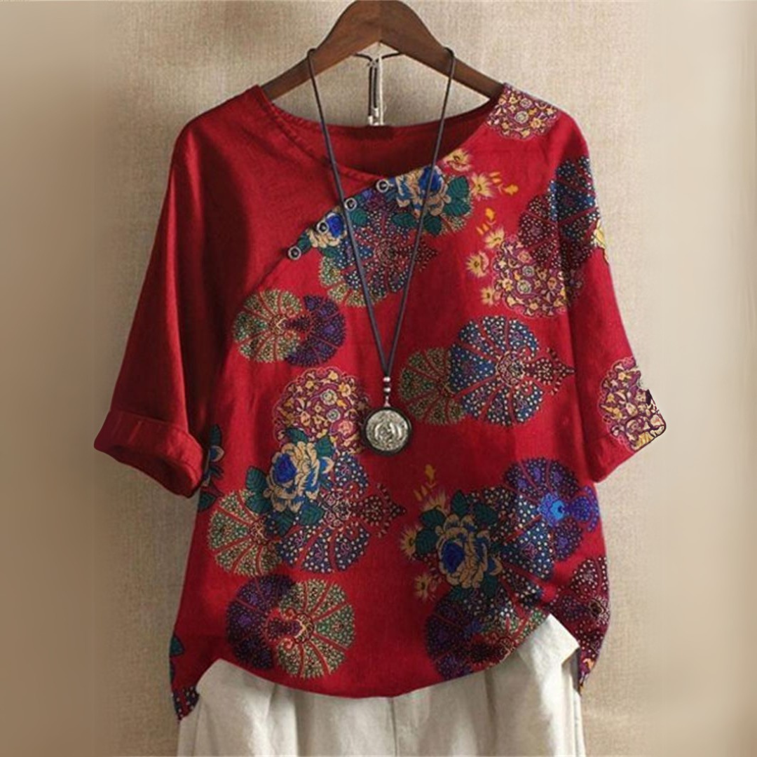Rotes Damenoberteil mit buntem Blumenmuster, asiatischem Design und Dreiviertelärmeln, hängend auf einem Holzkleiderbügel vor beigem Hintergrund.