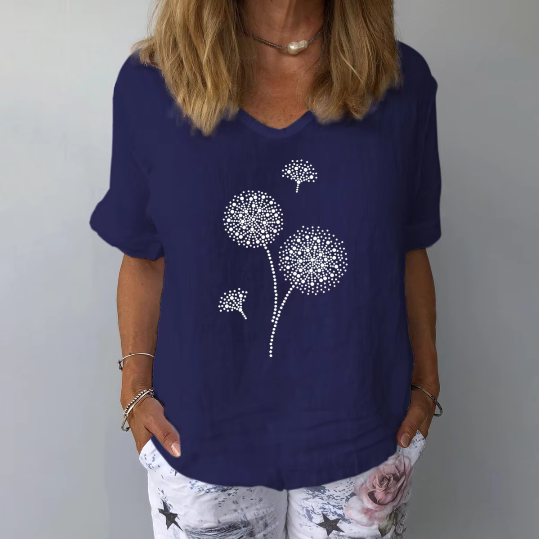 Frau trägt dunkelblaues T-Shirt mit Pusteblumen-Design, kombiniert mit weißer Hose mit Blumenmuster. Modetrend, Damenbekleidung, Sommeroutfit.