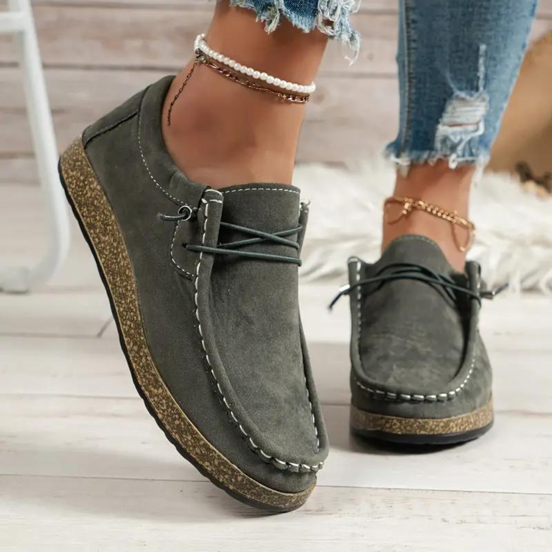Graue Damen-Mokassins aus Wildleder mit weißer Naht, getragen mit zerrissenen Jeans und Perlenfußkettchen. Bequeme Freizeitschuhe für den Alltag.