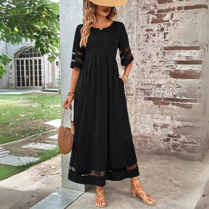 Frau in elegantem, schwarzem Maxikleid mit transparenten Ärmeln, Strohhut und runder Strohtasche, steht vor Backsteinmauer im Garten.