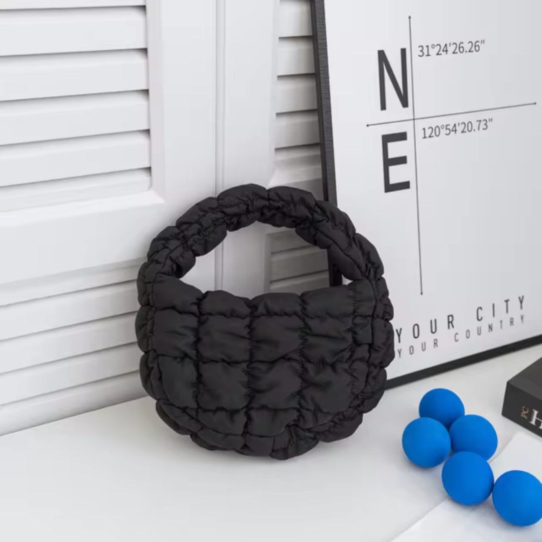 Schwarze gesteppte Handtasche auf weißem Tisch, umgeben von blauen Bällen und moderner Wanddekoration. Modische Accessoires, Inneneinrichtung.