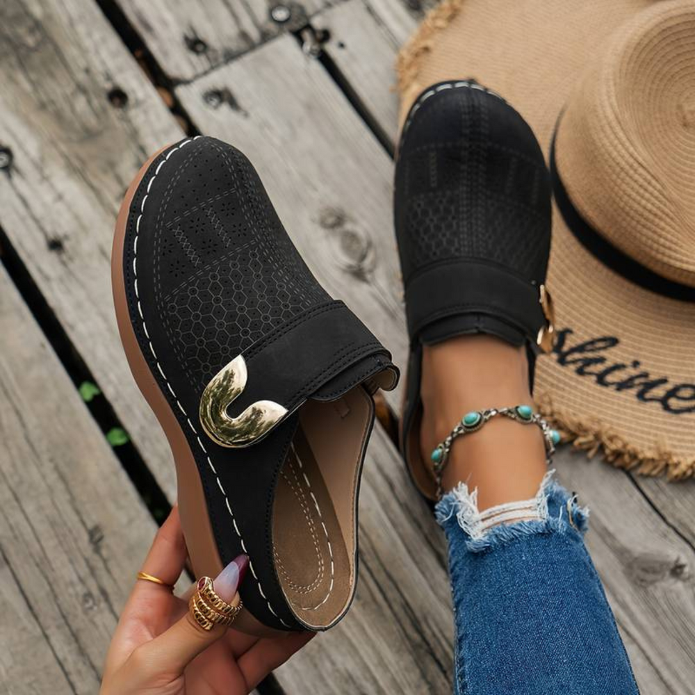Schwarze Damen-Loafer mit goldener Schnalle, getragen von Person in blauen Jeans, neben Strohhut auf Holzboden. Modische Schuhe, Sommerstil.