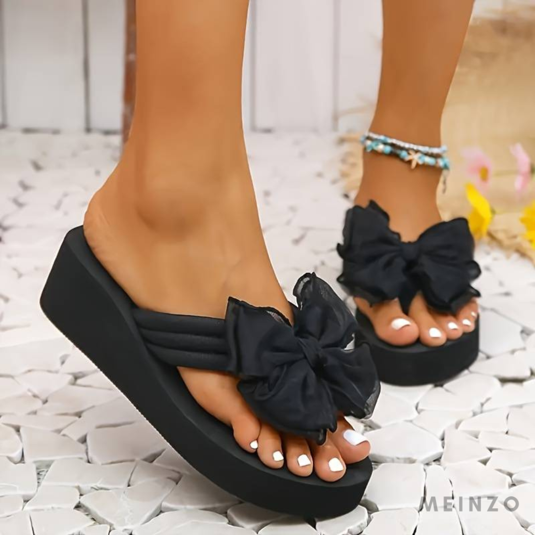 Schwarze Plateau-Sandalen mit großen Schleifen, getragen von einer Person mit weiß lackierten Zehennägeln auf weißen Kieselsteinen.
