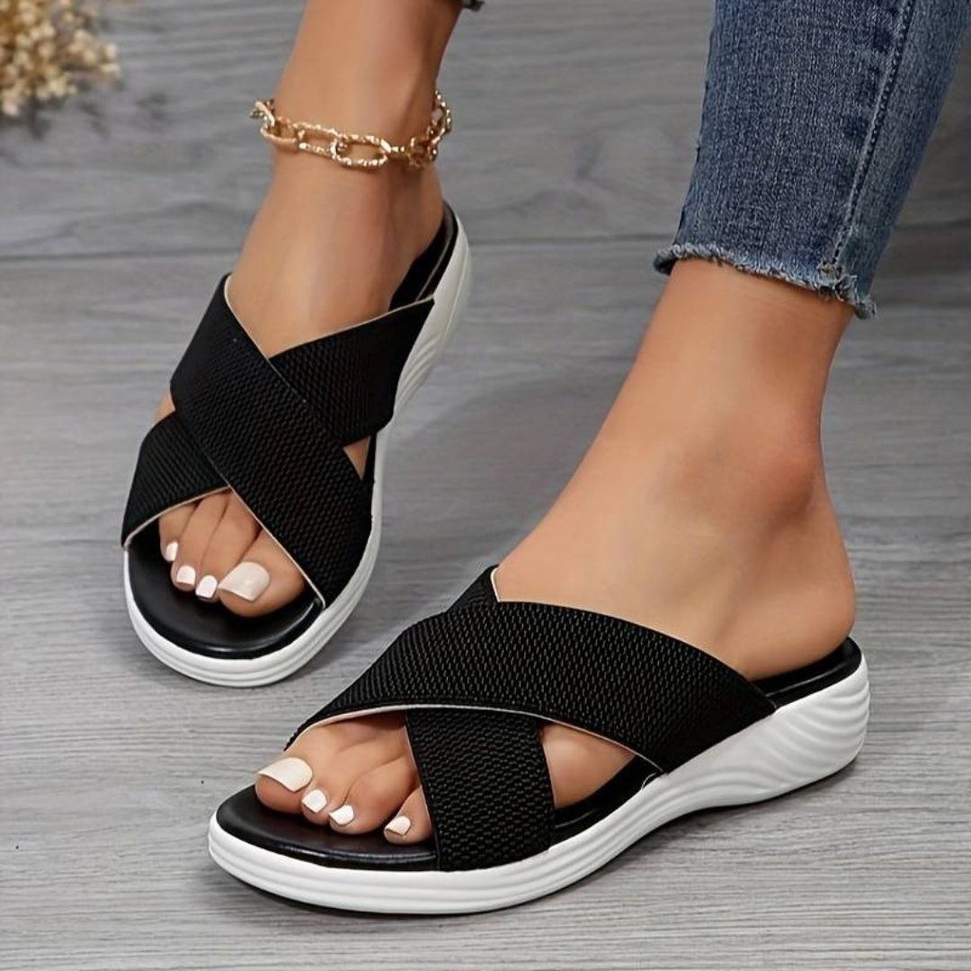 Damenfüße in schwarzen Sandalen mit Kreuzriemen und weißer Sohle, getragen mit blauen Jeans. Modische Sommerschuhe, bequem und stilvoll.