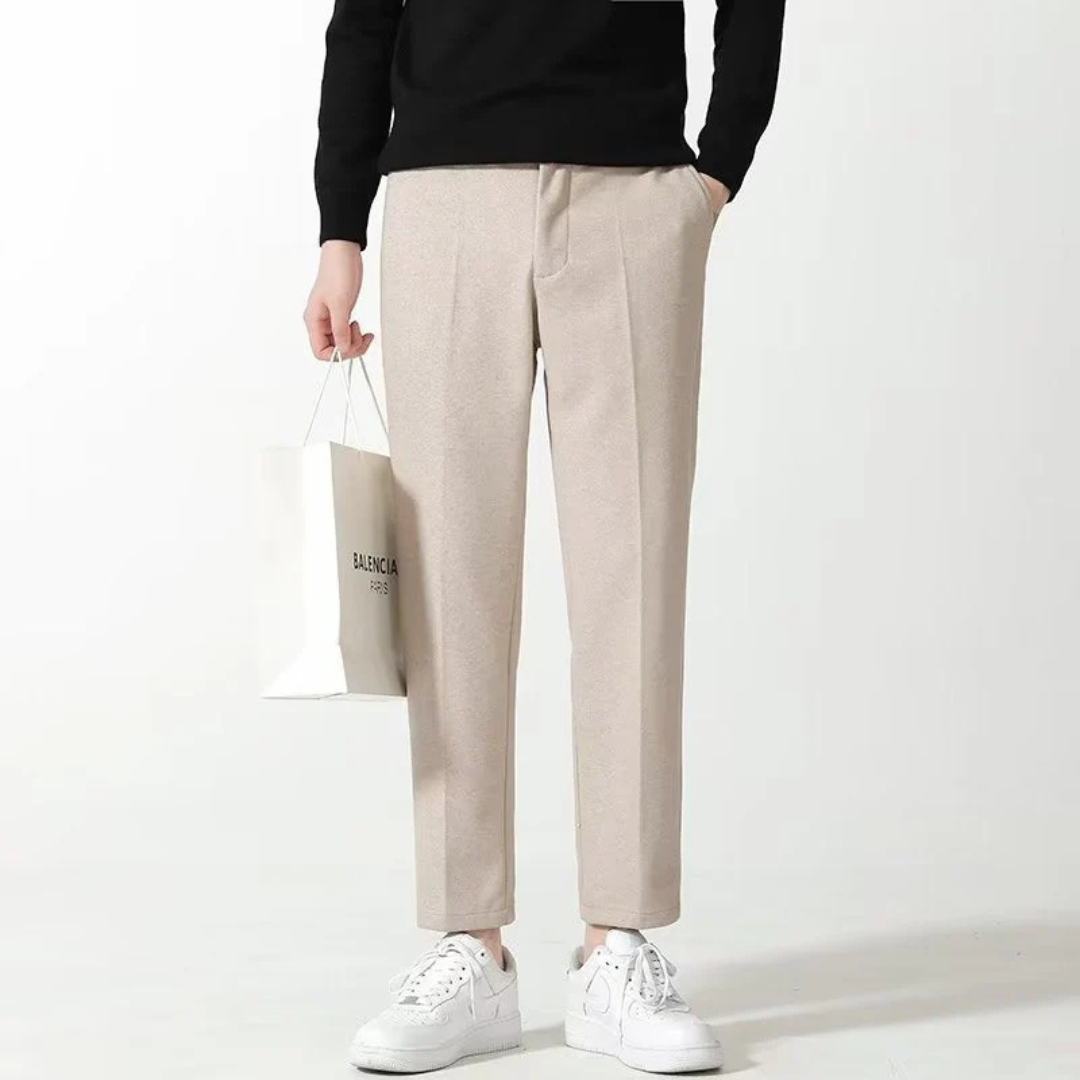 Beige Herrenhose, kombiniert mit weißen Sneakers und schwarzem Oberteil, Person hält Einkaufstasche. Elegante, moderne Herrenmode.