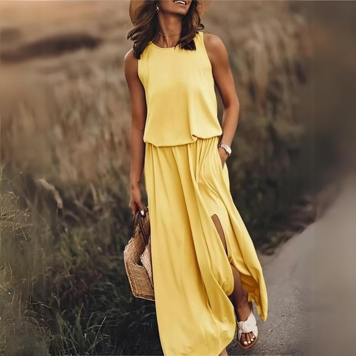 Frau in gelbem Maxikleid mit Schlitz, Strohhut und Korbtasche spaziert auf einem Weg. Sommermode, elegantes Outfit, Freizeitkleidung.