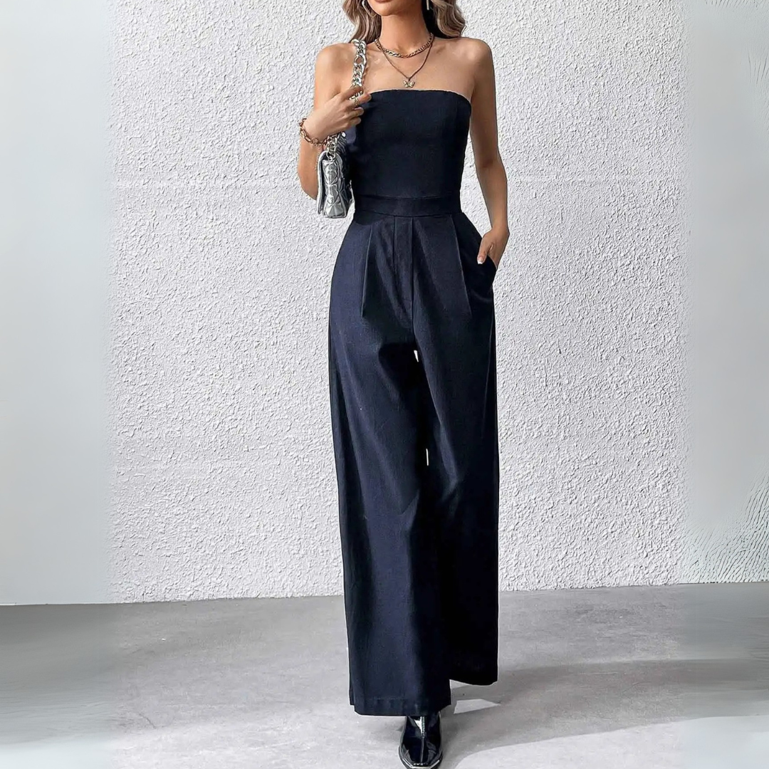 Frau in schwarzem, schulterfreiem Jumpsuit vor weißer Wand, modisch elegant, mit Handtasche und Schmuck, ideal für Abendmode und Events.