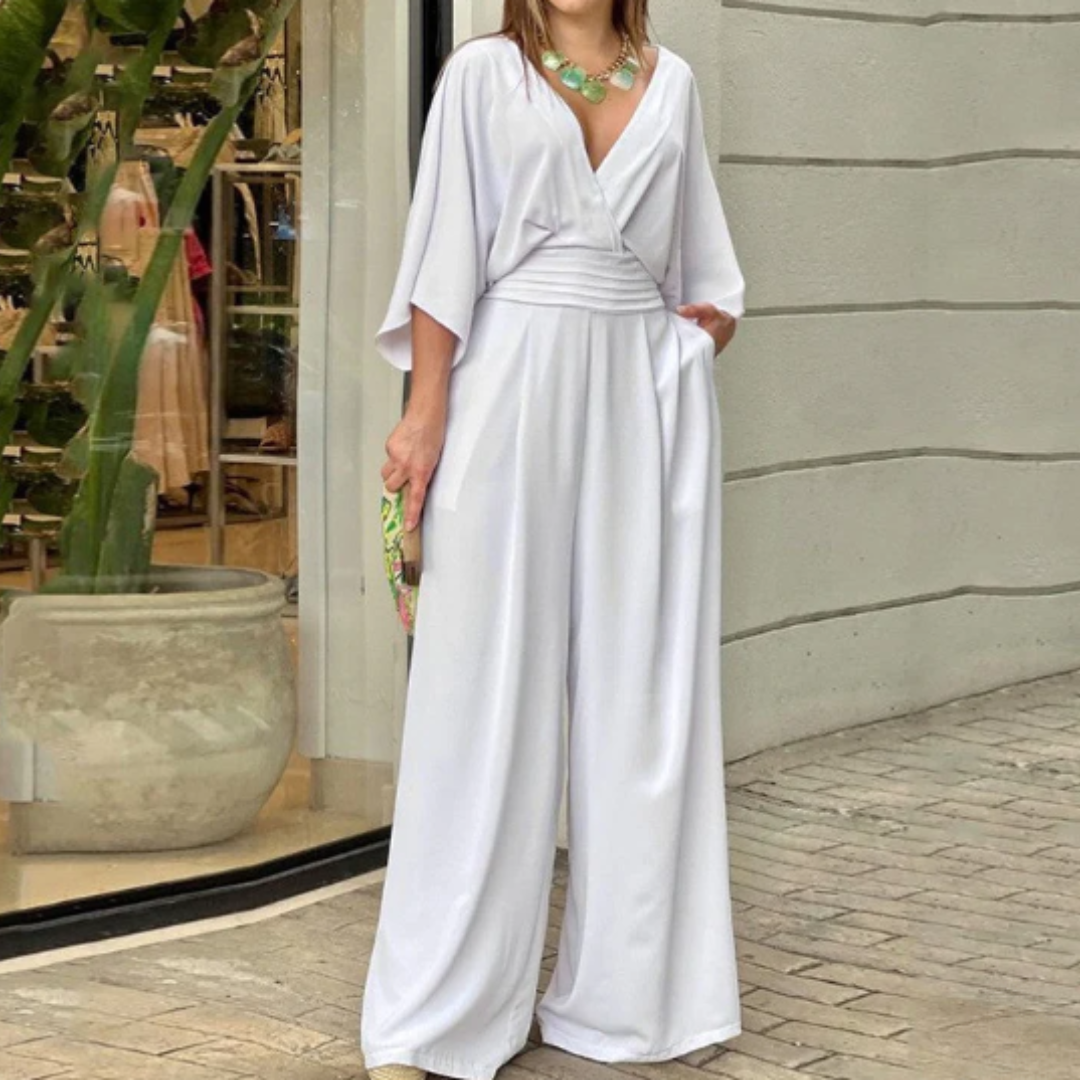 Frau in elegantem weißen Jumpsuit mit weitem Bein und V-Ausschnitt, steht vor einem Geschäft mit Pflanzen im Hintergrund. Fashion, Damenmode, Streetstyle.