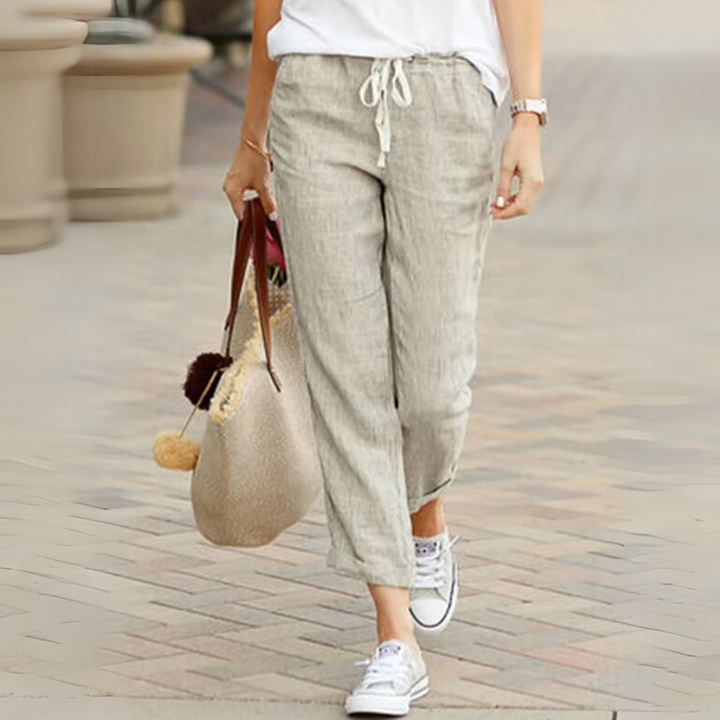 Dame in beigen Leinenhosen und weißen Sneakers, trägt eine beige Tasche mit Pompons. Perfektes Sommeroutfit für lässigen Stadtbummel.