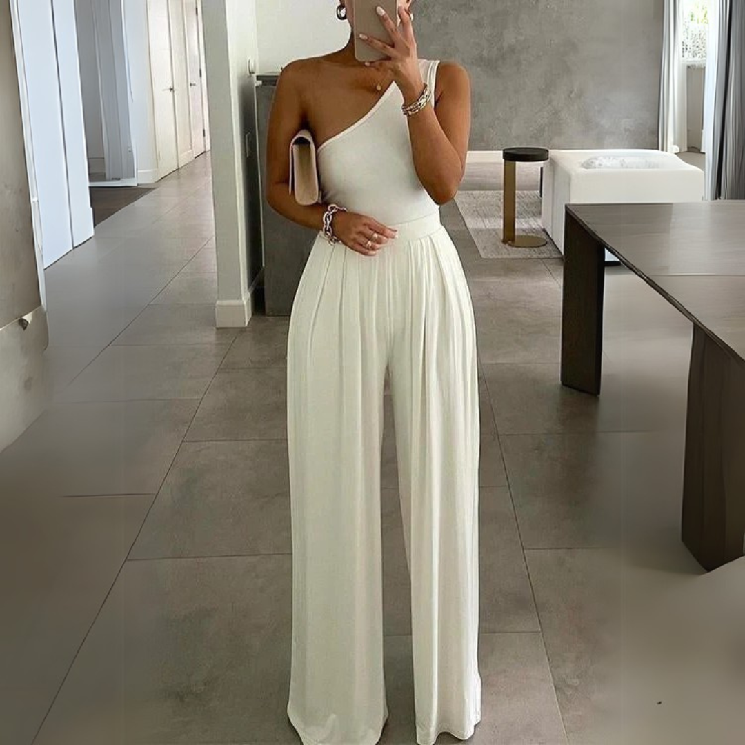 Frau in elegantem, weißem Ein-Schulter-Jumpsuit in modernem Wohnzimmer. Stilvolle Mode, schickes Design, trendige Damenbekleidung.