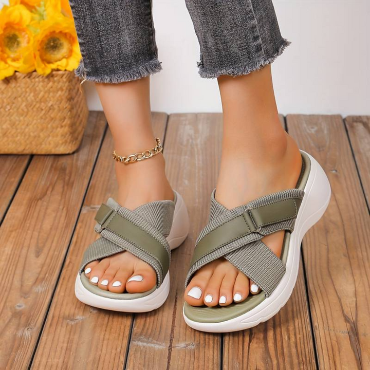 Damenfüße in grünen Sandalen mit Kreuzriemen auf Holzboden, Jeans hochgekrempelt, gelbe Blumen im Hintergrund. Bequeme Sommerschuhe, modisch.
