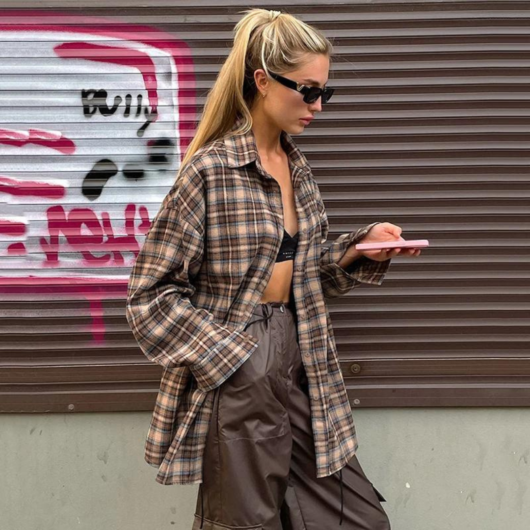 Frau in kariertem Hemd und brauner Hose, hält Smartphone. Hintergrund mit Graffiti. Streetstyle, Mode, urbane Kleidung, Sonnenbrille.