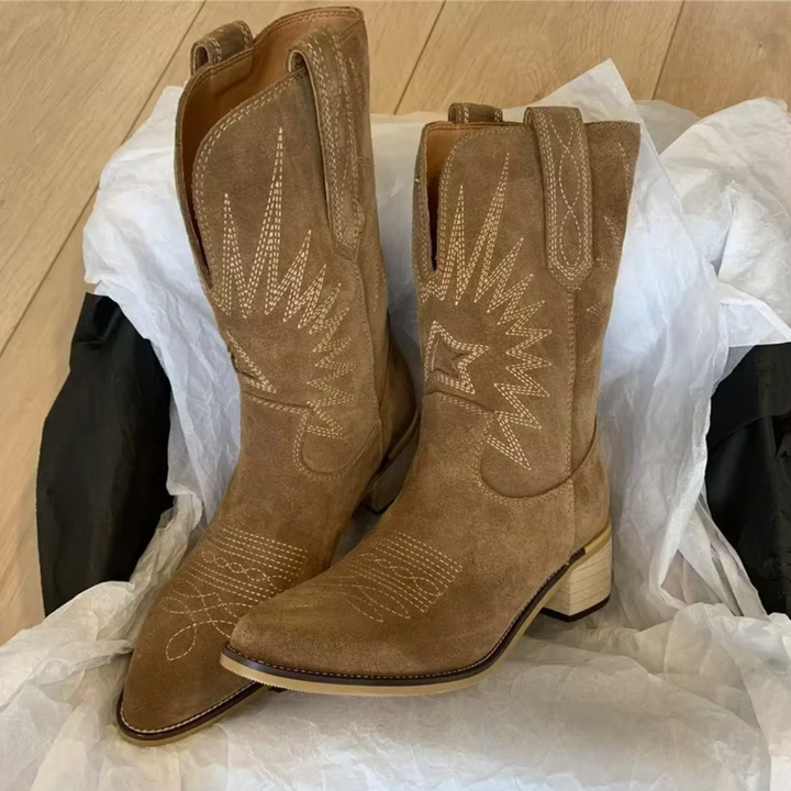 Braune Wildleder-Cowboystiefel mit Stickmuster, auf hellem Holzboden präsentiert. Westernstiefel, modisch, robust, ideal für Country-Look.