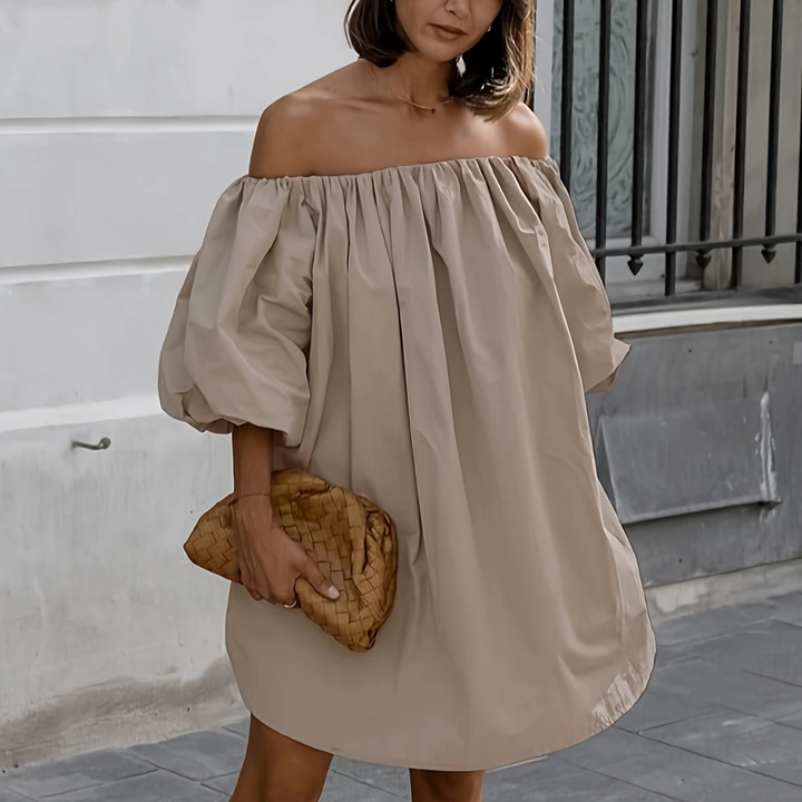 Frau in schulterfreiem, beigem Kleid mit Puffärmeln hält eine geflochtene Clutch. Elegante Sommermode, Streetstyle, Modeaccessoires.