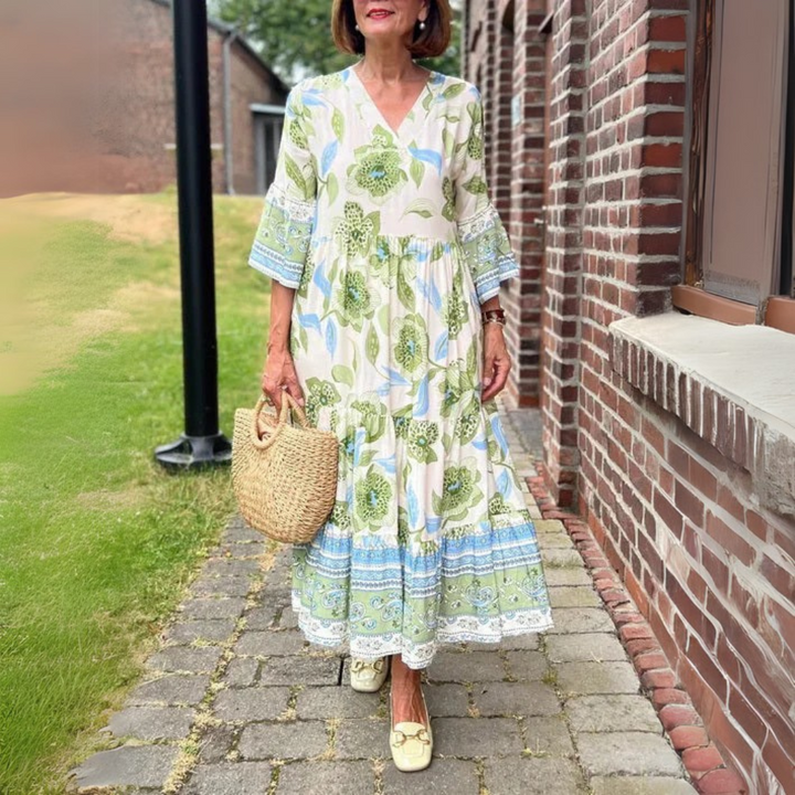 Frau in langem, geblümtem Sommerkleid mit Korbtasche, steht auf gepflastertem Weg neben Backsteinmauer. Elegante Mode, Sommeroutfit, Freizeitkleidung.