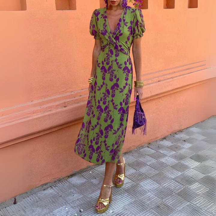 Frau in grünem, lila geblümtem Kleid, goldene Sandalen, lila Tasche, vor orangefarbener Wand. Sommermode, elegantes Outfit, Streetstyle.