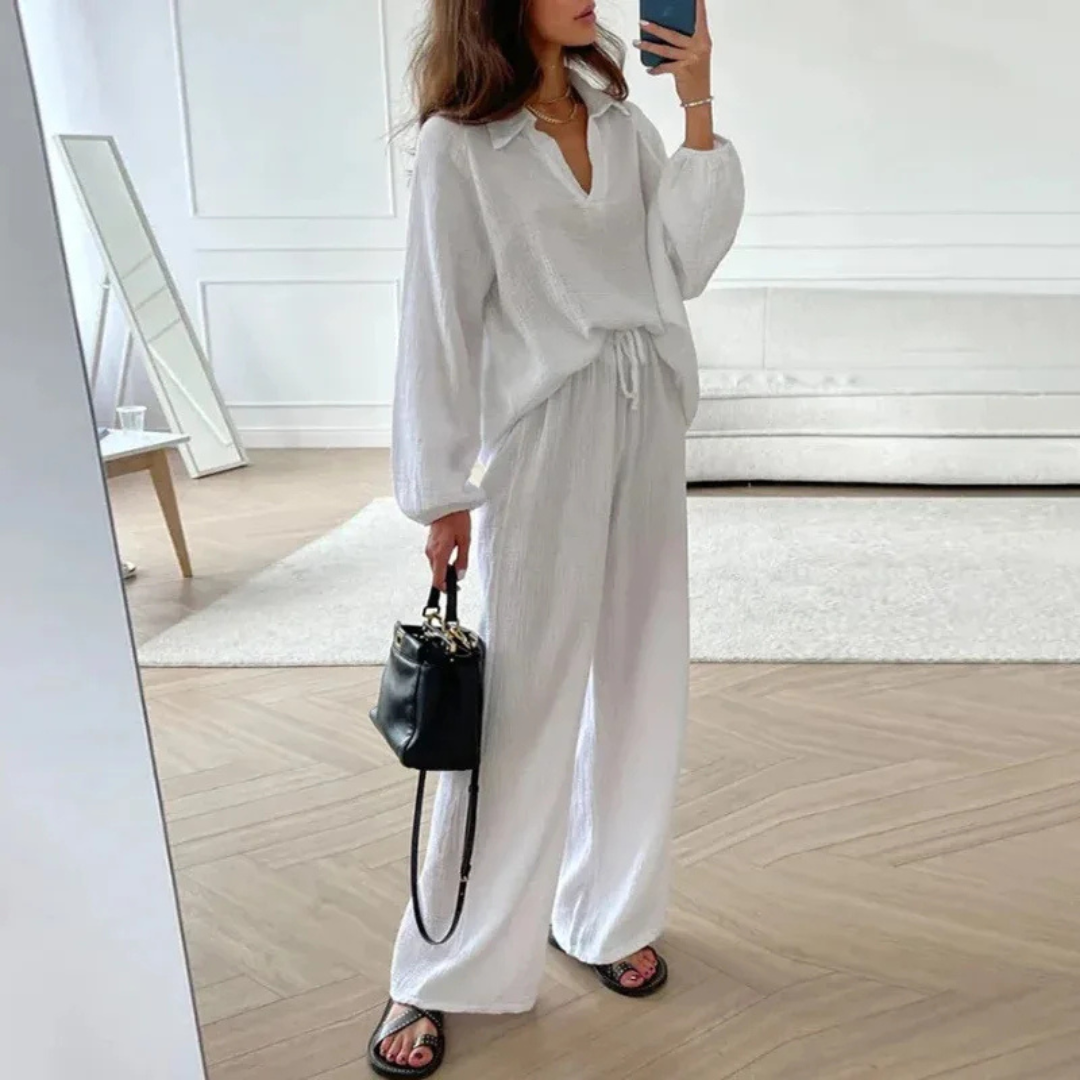 Frau in weißem, lockerem Leinenanzug mit schwarzer Handtasche und Sandalen, steht in modernem, minimalistischem Wohnzimmer mit Spiegel.