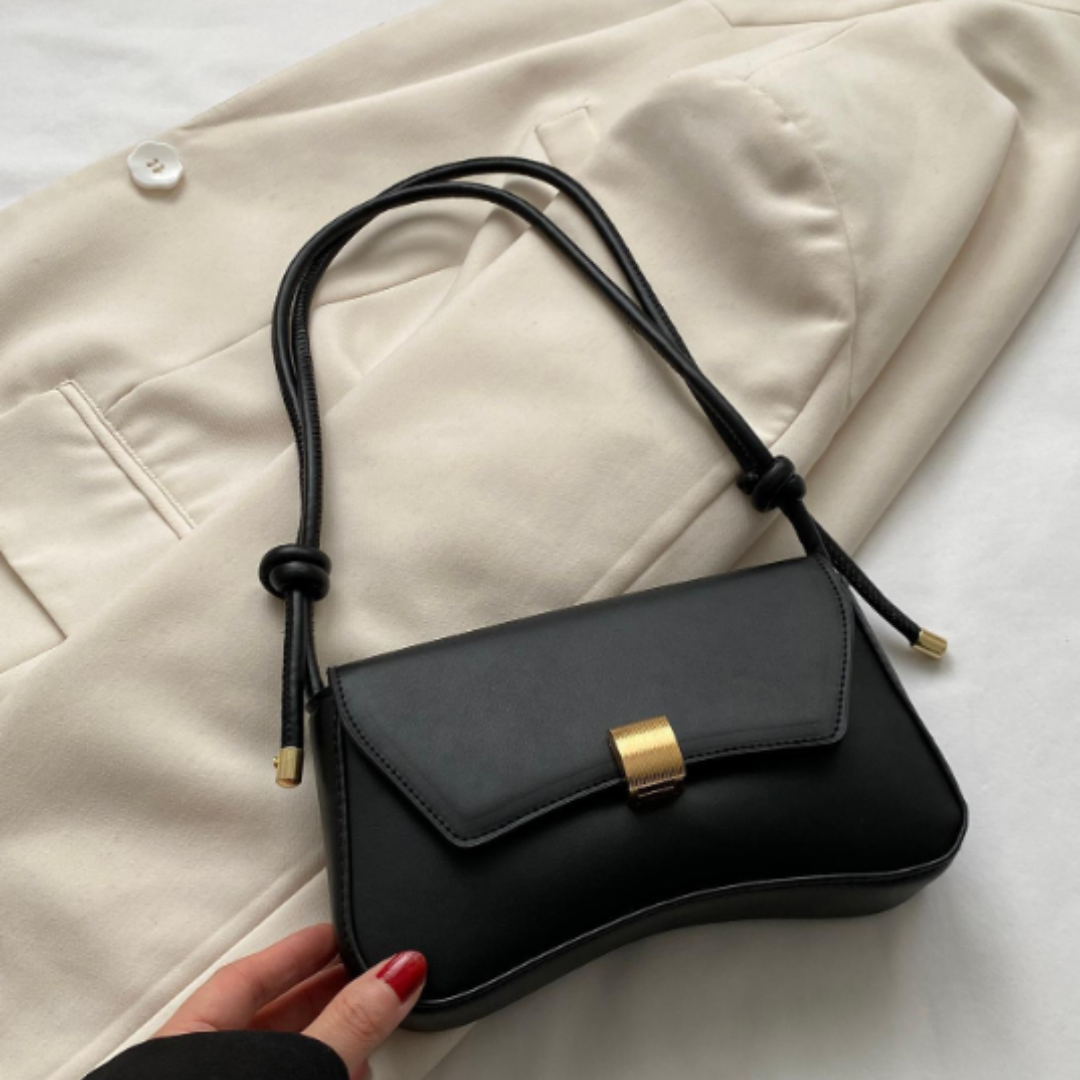 Schwarze Lederhandtasche mit goldener Schnalle auf beigem Blazer. Elegante Damenmode, stilvolles Accessoire, minimalistisches Design.