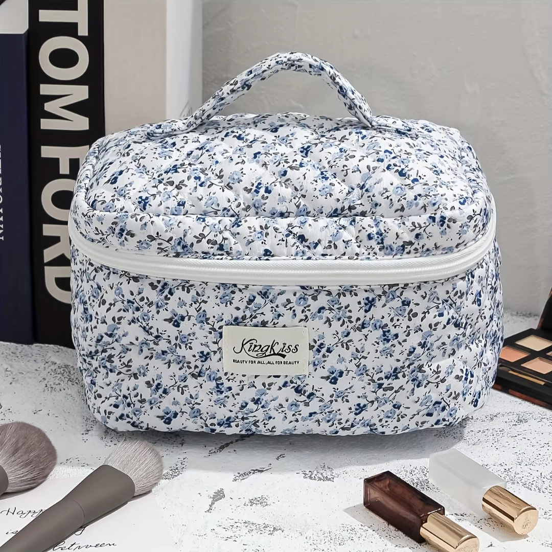 Blau-weiße Kosmetiktasche mit Blumenmuster, Reißverschluss und Tragegriff, umgeben von Make-up-Pinseln und Lippenstift auf einem Tisch.
