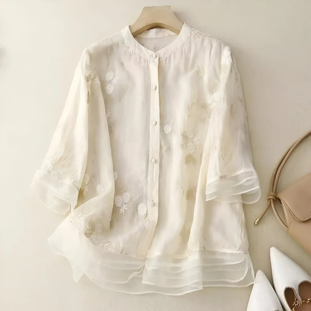 Weiße, transparente Bluse mit floralen Stickereien, Dreiviertelärmeln und Knopfleiste, elegant drapiert auf einem Kleiderbügel.