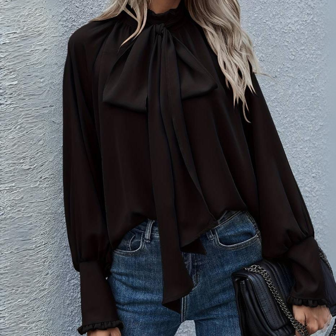 Blonde Frau in schwarzer Bluse mit Schleife und blauen Jeans vor grauer Wand. Modetrend, elegante Damenbekleidung, stilvolle Herbstmode.