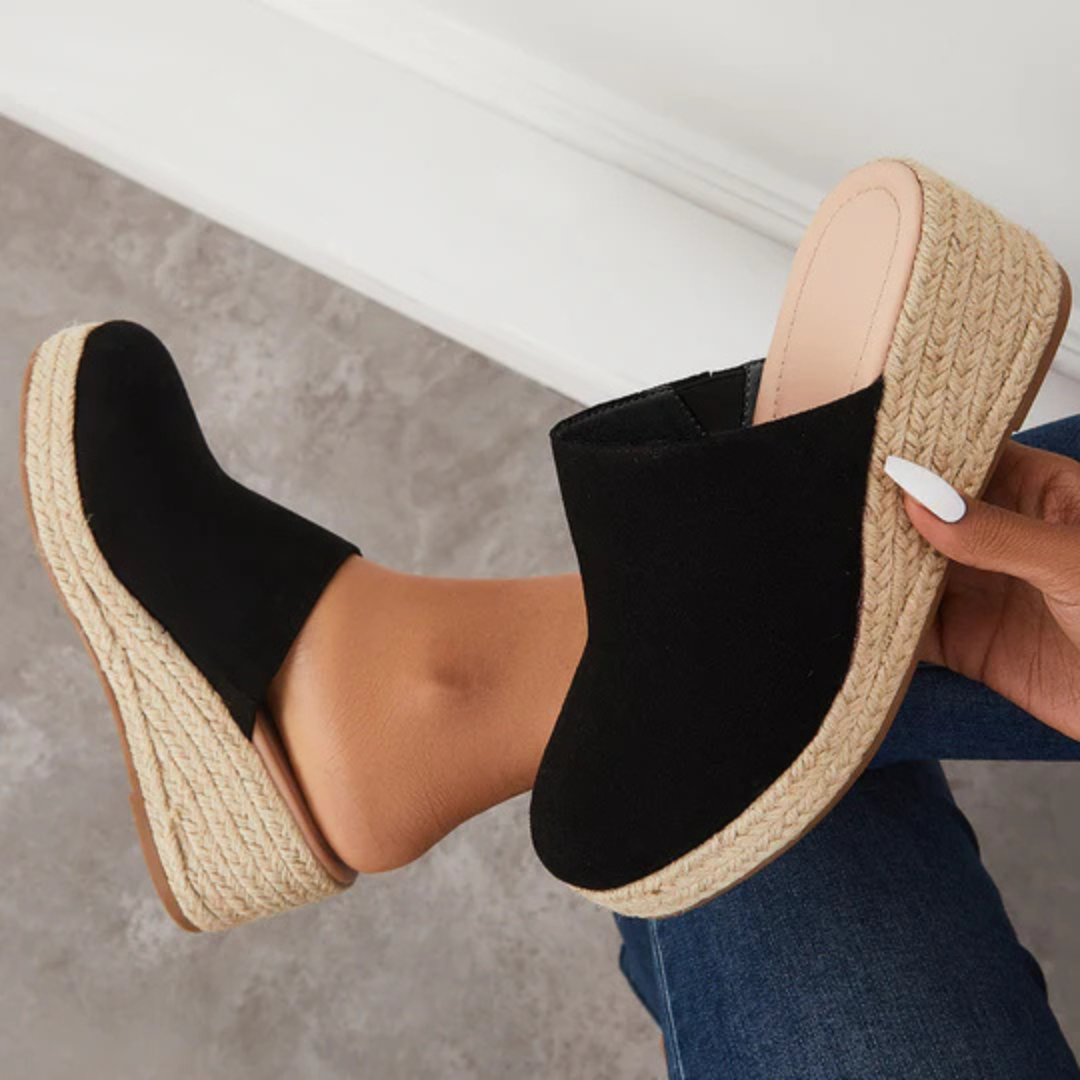 Schwarze Damen-Pantoletten mit Keilabsatz aus Jute, getragen von einer Person auf grauem Boden. Modische Sommerschuhe, bequem und stilvoll.