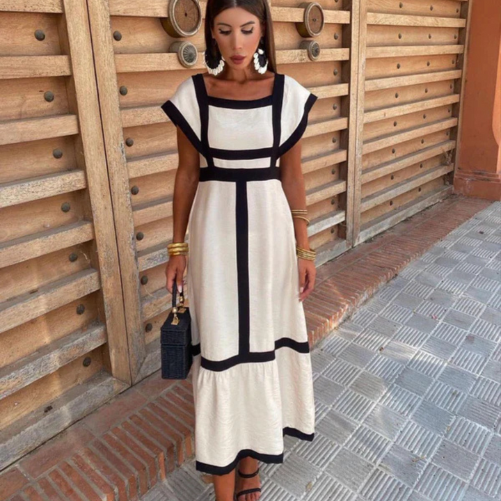 Frau in elegantem, weißem Kleid mit schwarzen Streifen, steht vor Holzwand. Sie trägt goldene Armreifen und Ohrringe, hält schwarze Handtasche.