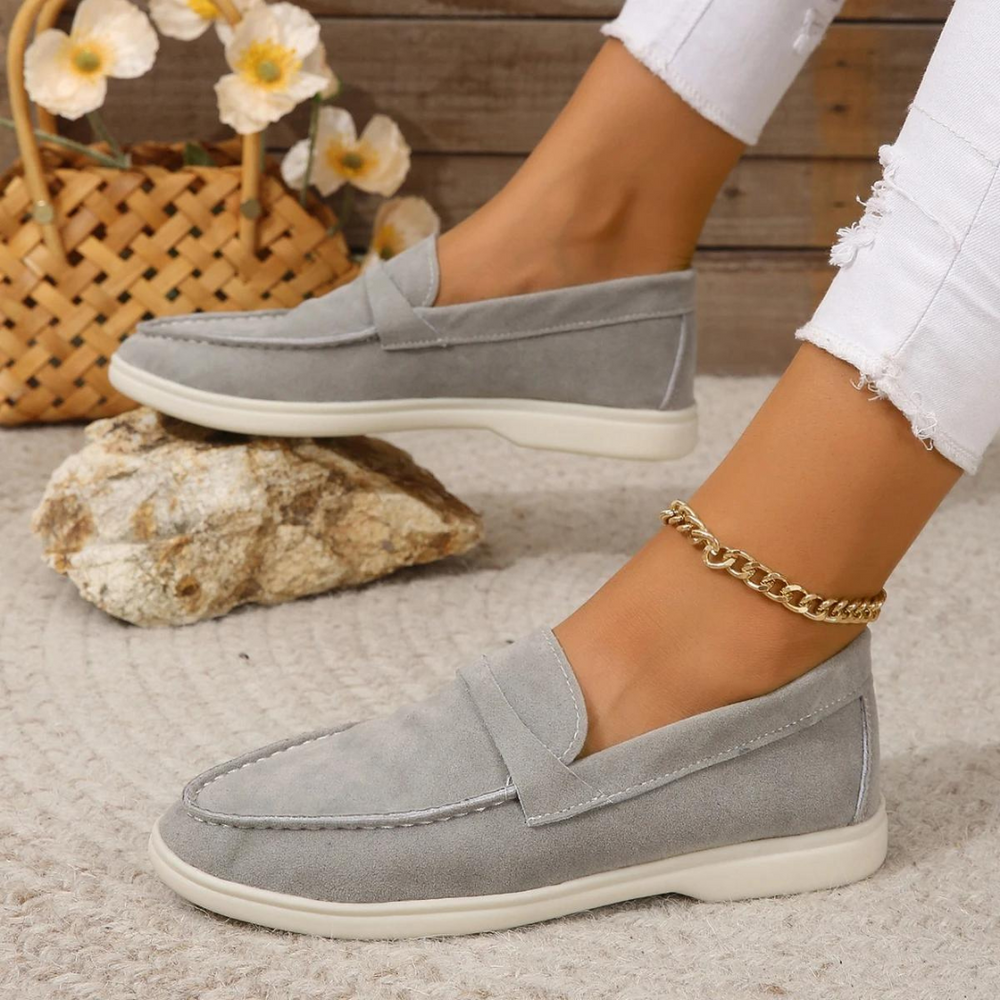 Graue Damen-Loafer aus Wildleder auf Steinboden, getragen mit weißer Jeans. Modische Schuhe, ideal für Freizeit und Sommer. Elegante Fußkettchen.