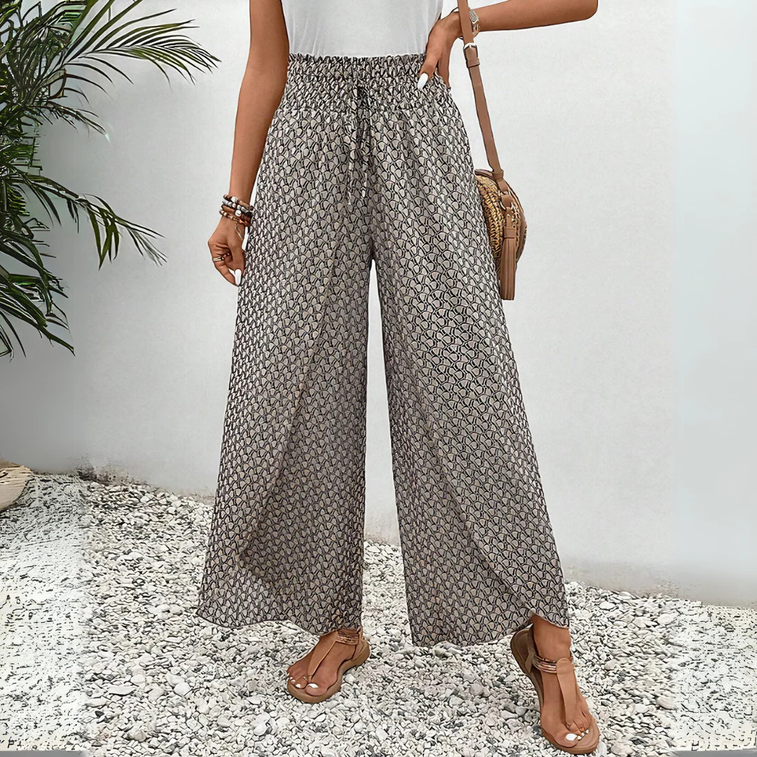 Frau in gemusterten Palazzo-Hosen, weißem Top und Sandalen, steht auf Kiesweg neben grüner Pflanze. Sommermode, lässiger Boho-Stil.