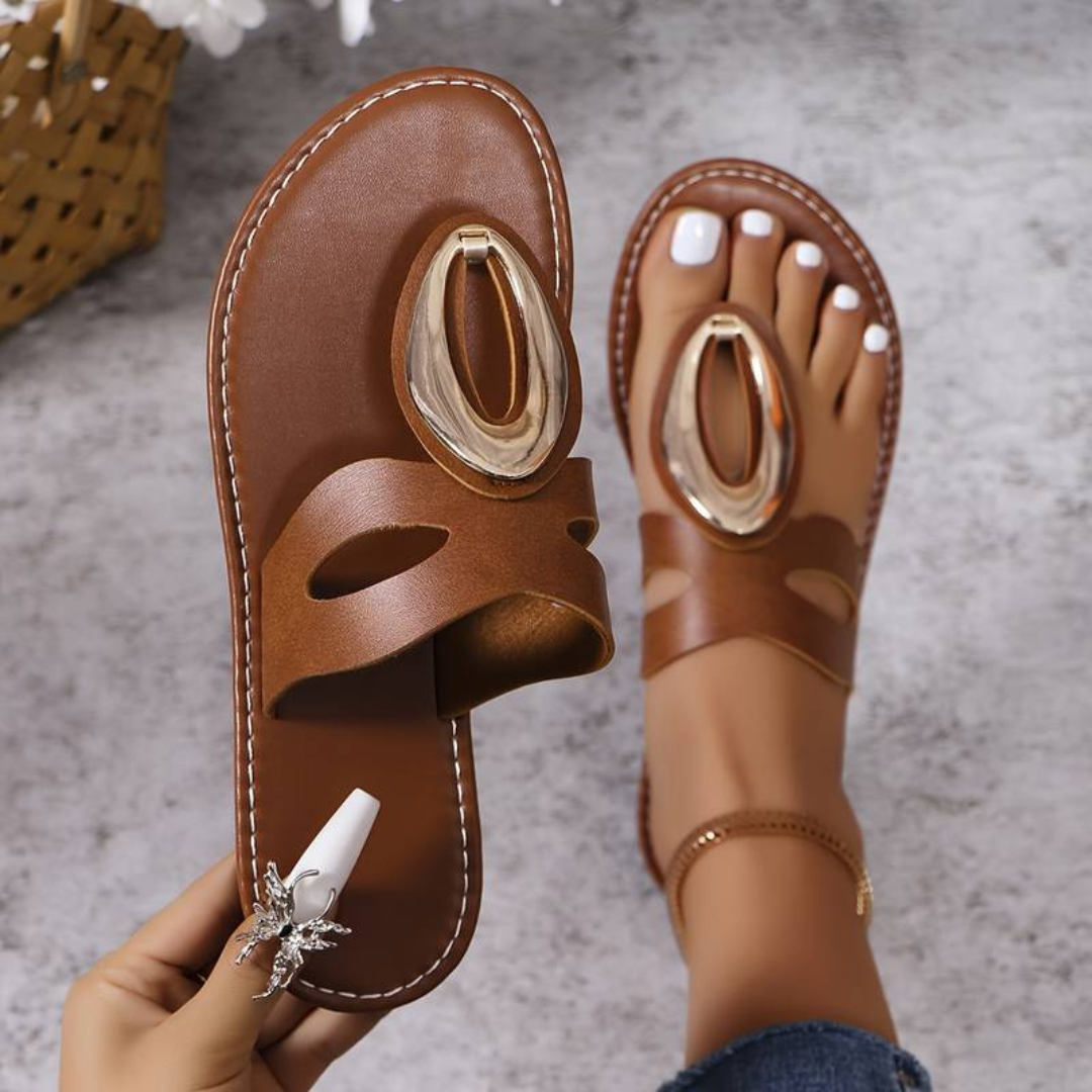Braune Ledersandalen mit goldener Verzierung, getragen von einer Person mit weiß lackierten Zehennägeln. Modische Sommerschuhe auf grauem Hintergrund.