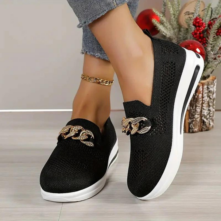 Schwarze Damen-Sneaker mit goldener Kettendekoration, weißer Sohle und Strickdesign, getragen mit Jeans. Modische Freizeitschuhe für Damen.