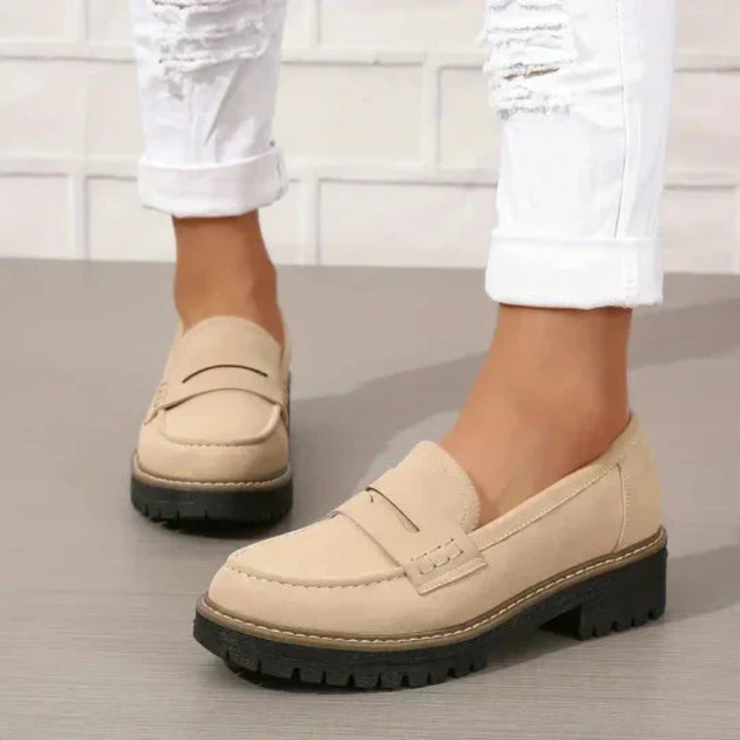 Beige Damen-Loafer mit dicker Sohle, getragen mit weißen, zerrissenen Jeans vor einer weißen Ziegelwand. Modische Schuhe, bequeme Damenmode.