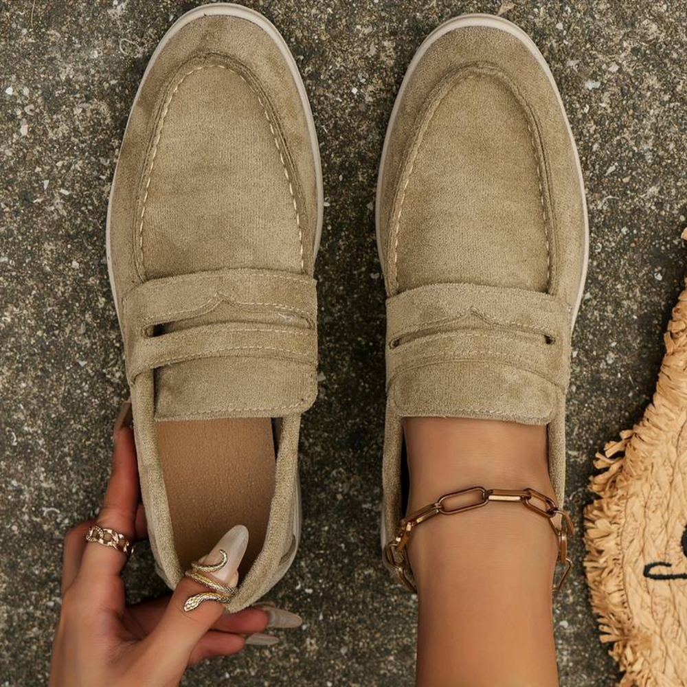 Damenfüße in beigen Wildleder-Loafern auf grauem Boden, modische Schuhe, elegantes Design, goldene Fußkette, stilvolle Damenmode.