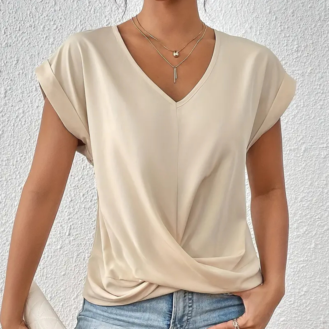 Frau trägt beiges, kurzärmeliges V-Ausschnitt-Shirt mit Knoten-Detail, kombiniert mit blauer Jeans und goldener Halskette vor weißer Wand.