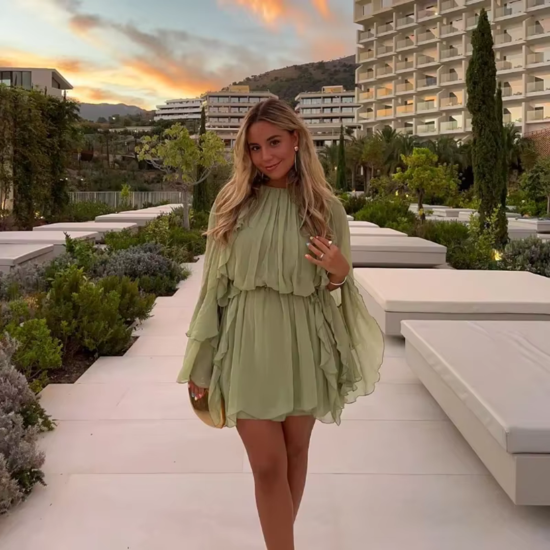 Eine Frau in einem fließenden, hellgrünen Kleid posiert vor einem modernen Hotelgebäude bei Sonnenuntergang. Elegante Mode, Sommerabend, stilvolle Umgebung.