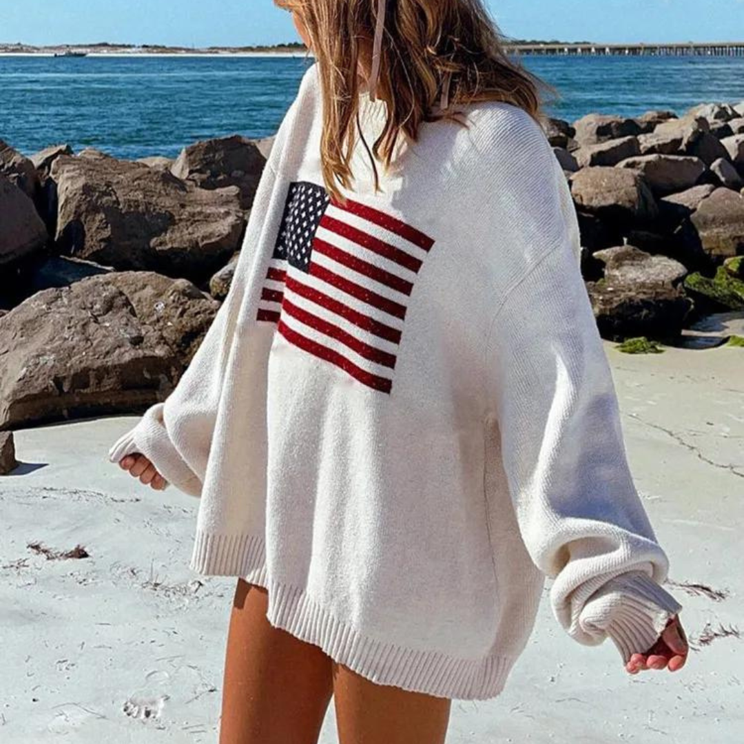 Frau am Strand in übergroßem weißen Pullover mit USA-Flagge, Meer und Felsen im Hintergrund. Sommermode, Strandkleidung, lässiger Stil.