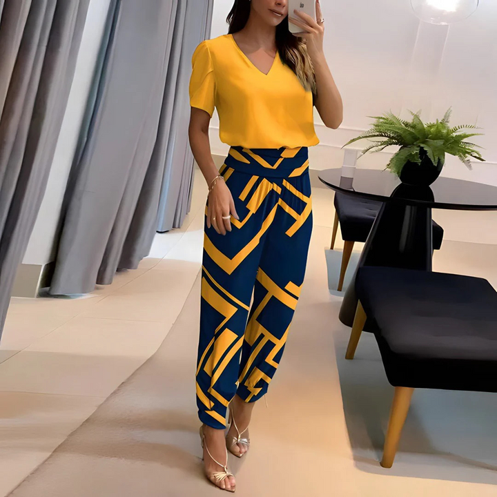 Frau in gelber Bluse und geometrisch gemusterter blauer Hose, steht in modernem Wohnzimmer mit Pflanzen und stilvollen Möbeln. Fashion, Mode, Inneneinrichtung.