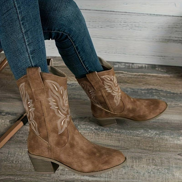 Damen Cowboy-Stiefel aus braunem Wildleder mit Stickerei, kombiniert mit blauen Jeans. Modische Westernstiefel auf Holzboden.