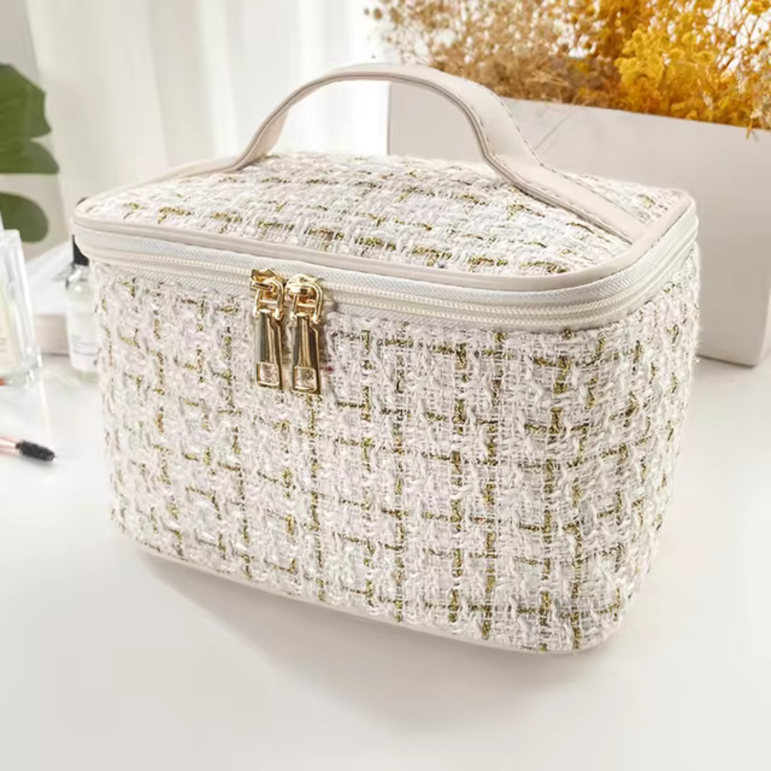 Luxuriöse, cremefarbene Kosmetiktasche aus Tweed mit goldenen Reißverschlüssen auf weißem Tisch, umgeben von Pflanzen und Make-up-Accessoires.