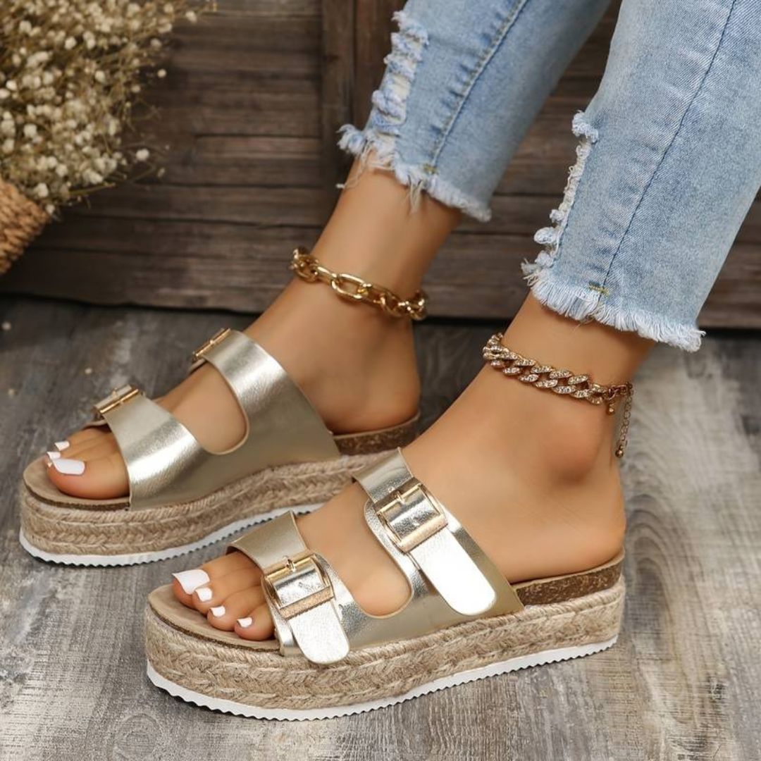 Goldene Plateau-Sandalen mit Schnallen an Frauenfüßen, kombiniert mit zerrissenen Jeans und Fußkettchen, auf Holzboden. Modetrend Sommer 2023.