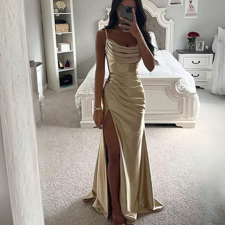 Frau in elegantem, goldfarbenem Abendkleid mit hohem Beinschlitz, steht in stilvollem Schlafzimmer mit weißem Bett und Dekoration im Hintergrund.
