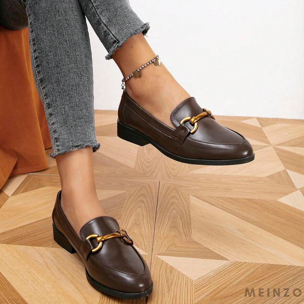 Braune Damen-Loafer mit goldener Schnalle, getragen mit grauen Jeans auf Holzfußboden. Elegante, modische Schuhe für stilvolle Outfits.