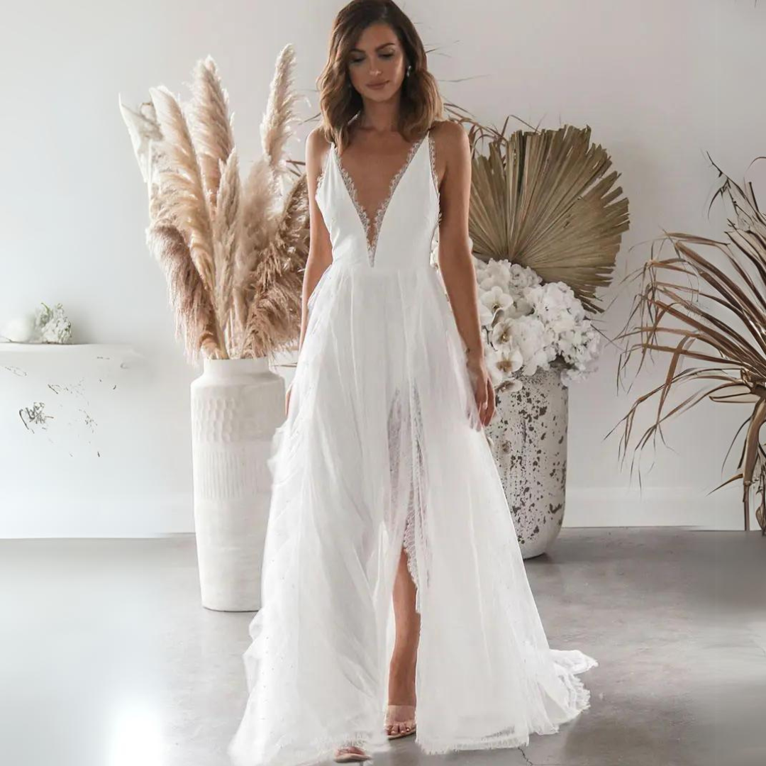 Braut in elegantem, weißem Hochzeitskleid mit tiefem V-Ausschnitt, umgeben von Pampasgras und Trockenblumen in modernem, minimalistischem Raum.