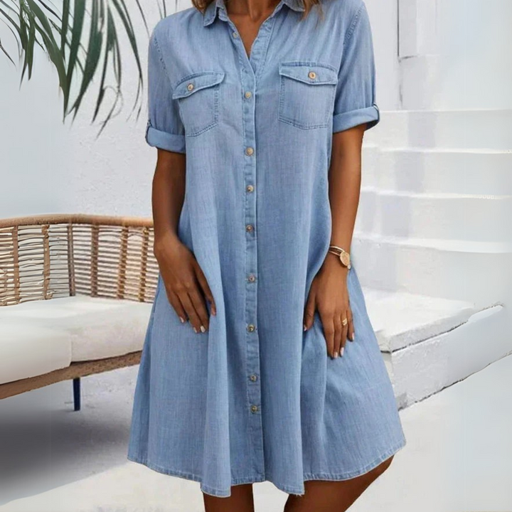 Frau in hellblauem Jeanskleid mit Knopfleiste, stehend vor weißer Wand und Rattanmöbel. Sommermode, Freizeitkleid, Denim-Look.