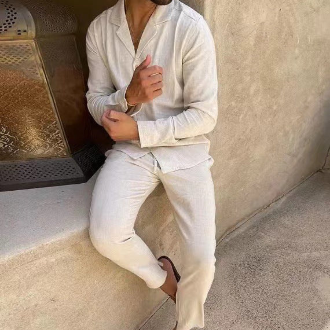 Mann in beiger Leinenkleidung lehnt an einer Wand, trägt Sandalen. Sommermode, lässiger Stil, entspannte Atmosphäre.